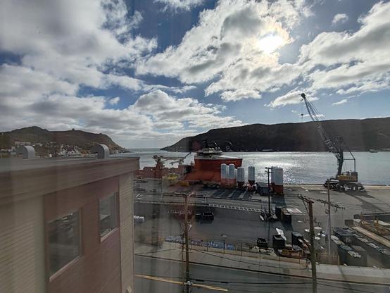Picture of a natural harbour, looking out to the narrow entrance set between two rocky hillsides. The sky is covered in streaky white clouds, through which the sun is trying to pierce. In the foreground, a supply ship is moored, equipped with an icebreaking prow and elevated control deck forward, the aft having a lower, flat deck and crane.