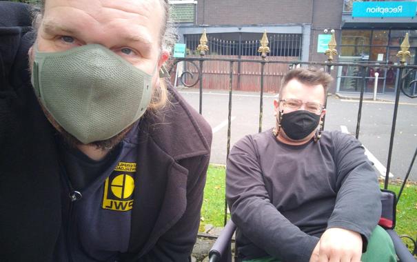 E is sitting in a folding camp chair in front of some railings outside a hotel being used to house refugees. I'm squatting next to him. We are white guys wearing masks and dark clothing.