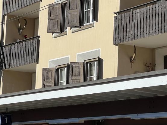 Das Bild zeigt eine Gebäudefassade mit zwei Balkonen mit Holzgeländern. Ein Balkon zeigt Tierhörner, die an der Wand angebracht sind, neben einem roten Ornament. Fenster unter den Balkonen haben Holzläden, die offen sind. Ein Schild mit der Nummer "1" ist sichtbar