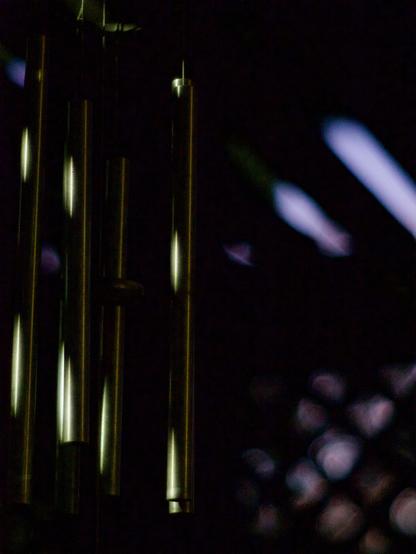 A set of wind chimes reflects the golden-orange light of a streetlight, contrasting against streaks of blue evening sky peeking through slats in the background.