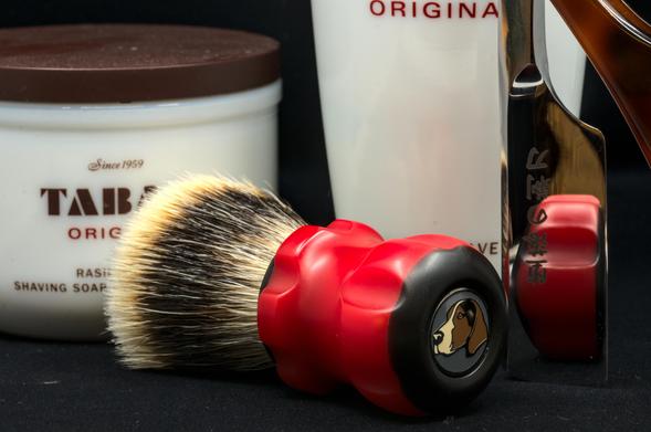 (Le Chat Mistral description)

This image shows a collection of shaving items on a dark background. In the foreground, there is a shaving brush with a vibrant red and black handle, featuring a golden emblem of a dog’s head. The bristles are a mix of light and dark tones.

Behind the brush, there is a white container of Tabac Original shaving soap, labelled "Since 1959." To the right of the brush, there is a straight razor with a metallic blade and a red-scaled handle, partially visible.