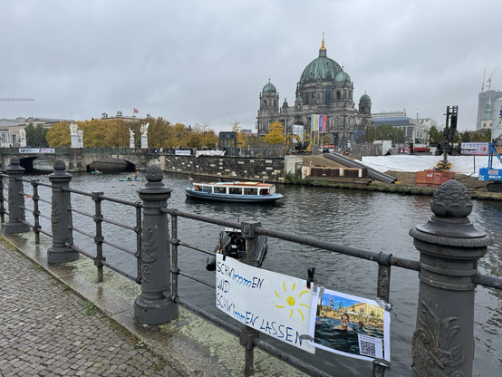 Blick über die Spree in Berlin. Im Vordergrund ein Geländer, an dem ein Schild mit der Aufschrift „schwimmen ins schwimmen lassen“ angebracht ist. Dahinter die sie mit ein paar Booten vielen Schwimmern. Im Hintergrund herbstgelbe Bäume und der Berliner Dom.