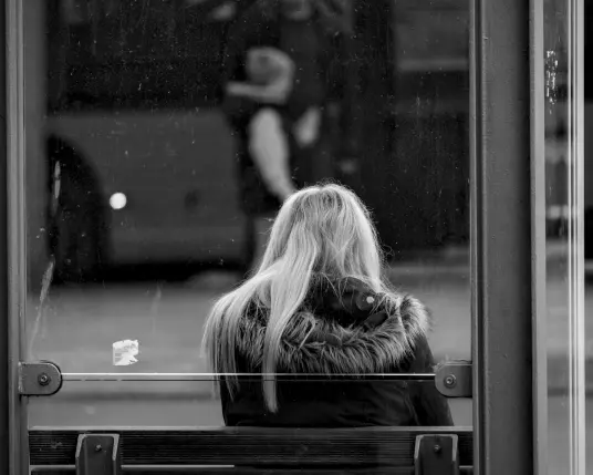 Eine Frau mit langen blonden Haaren sitzt auf der Bank eines Bushäuschens. Man sieht sie von hinten durch die Glasscheibe des Bushäuschen. Vor ihr steht ein Bus, in den soeben eine Person einsteigt.
.
A woman with long blonde hair is sitting on the bench of a bus shelter. She can be seen from behind through the glass pane of the bus shelter. In front of her is a bus, which a person is just getting on.
.