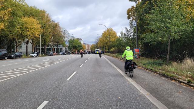 Querformat in Farbe. Blick die Hauptstraße 'An der Verbindungsbahn' entlang. Fünf oder sechs Spuren für den KFZ Verkehr, eine Parkspur, sowie ein ca 1,20 bis 1,50 m schmaler Streifen für den Radverkehr. Links und rechts stehen herbstlich verfärbte Laubbäume. Die Fahrbahn ist, bis auf einige Fahrradfahrer*innen leer, im Hintergrund stehen aber viele Menschen mit ihren Rädern über die gesamte Breite. Der Himmel ist wolkig.