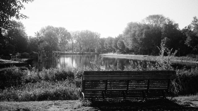 Black & white photo of a bench overlooking Noordhollandsch Kanaal, the Florapark, and on the left a little boat.