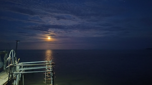 photo de la lune au-dessus d'un lagon.  Au premier plan, un ponton et un portique à bateau sont éclairés par un lampadaire. La lune, légèrement masquée par des nuages se reflète sur l'eau calme du lagon