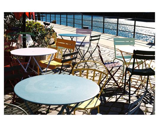 An outdoor seating area by the water in Hafencity, Hamburg, drenched in bright sunlight. Several round, brightly colored bistro tables and folding chairs are haphazardly arranged on a cobblestone pavement. Dominant colors are pastel blue, mint green, mustard yellow, and deep purple-red. The sun-dappled blue water is visible beyond a dark metal railing, and sharp, geometric shadows are cast across the scene.