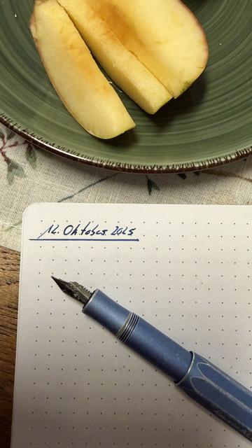 Ein Foto zeigt ein Notizbuch mit einem blauen Füllfederhalter darauf. Das Notizbuch ist geöffnet, mit Punkten über die Seite verteilt. Der Füllfederhalter ist horizontal angeordnet und seine Spitze befindet sich auf der Seite. Im Hintergrund ist ein grüner Teller mit einem halben Obststück darauf zu sehen. Auf dem Notizbuch ist Text geschrieben. Der Text lautet "12. Oktober 2015".