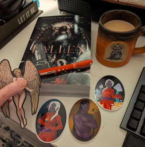 Author swag on a white desk: a paperback of Fallen: Darkness Ascending, 4 stickers featuring angel, dark elf, half-dragon. An orange and black mug with creamed coffee featuring a small jack-o-lantern Halloween spirit.