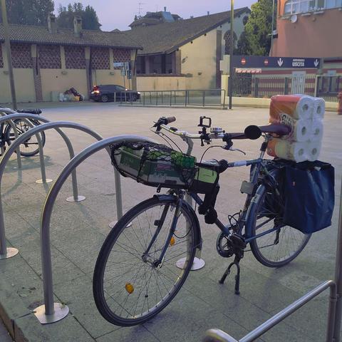 Una bici blu da uomo in una rastrelliera. La bici è carica: due borse laterali blu piene, una confezione di scottex e una cassetta di frutta nel portapacchi anteriore.

È sera, dietro si vede una cascina