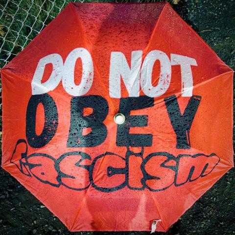 The bright-red top of an open umbrella resting on the ground reads, "DO NOT | OBEY | FASCISM," in large, blocky, black-and-white lettering.