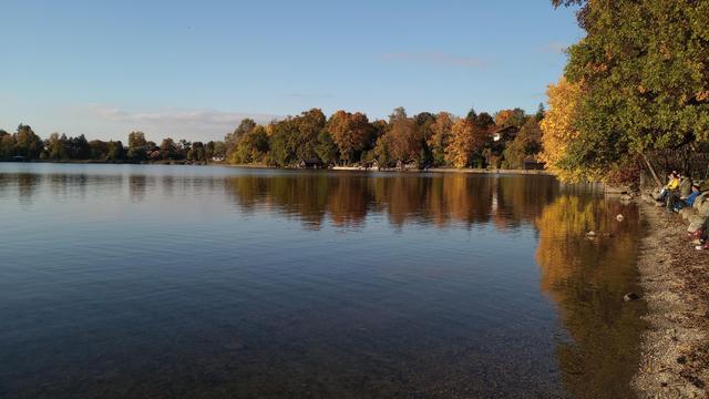 Blick auf einen See. Am Ufer spiegeln sich die herbstlich gefärbten Bäume im Wasser.