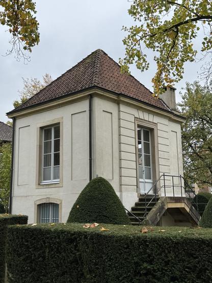 Ein kleines Gebäude mit quadratischem Grundriss aus der Zeit des Barock. Exakt geschnittene Sträucher, einer in Zuckerhutform