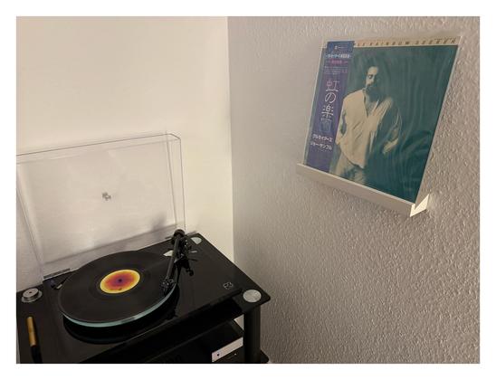 Auf einem schwarzen Plattenspieler läuft eine schwarze Schallplatte. Rechts an der Wand ist ein kleines weißes Regalbrett, darauf steht das Cover. Ein Bild des Künstlers mit geschlossenen Augen. Links ist eine Banderole mit japanischen Schriftzeichen.