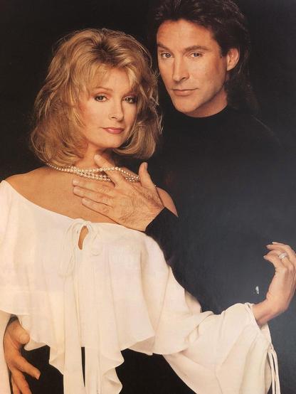 Professionally shot 1994 portrait of Deidre Hall and Drake Hogestyn, known for their roles as Marlena Evans and John Black on Days of Our Lives. Deidre wears an off-the-shoulder cream top with a dramatic hanging ruffle and a string of pearls. Her shoulder-length hair is softly layered in shades of blonde and light brown, styled with volume and elegance. Drake stands beside her in a dark sweater, gently lifting her pearls with one finger in an intimate gesture. Both look directly at the camera with composed, emotionally charged expressions. The background is solid black, enhancing the romantic and dramatic tone. The image evokes the intensity of the Maison Blanche storyline, where love, memory, and identity collided in classic 90s soap fashion.