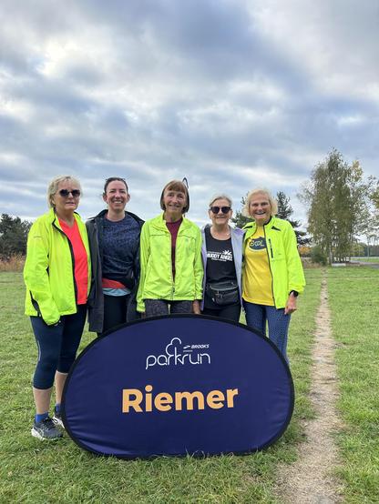 Group of people posing for a photo with the Riemer parkrun pop-up sign in front. The setting is a park.