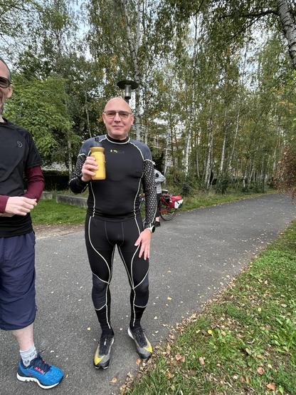 A man wearing tight running clothing, holding golden bottle. In the background many trees are visible.