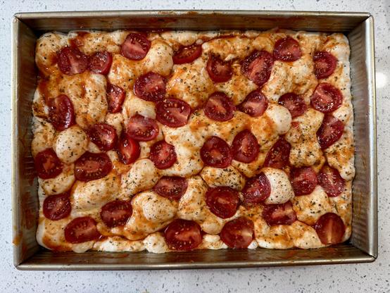 Focaccia dough in a steel baking tray with a thin layer of a red tomato sauce on top. Cherry tomato halves are studded throughout the dough, cut side up. Some flakes of salt and Italian herb seasoning can be seen.