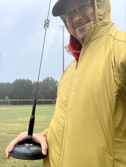 Man in a yellow rain jacket holding a black magnetic mount whip antenna in the rain.