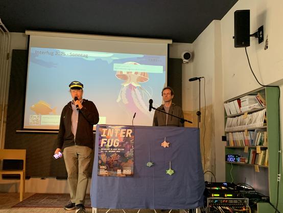 Bild zeigt die Bühne im Lokomov, Beamer blendet nautisches Motiv der Interfug 25 ein, links Mann mit Kapitänsmütze, der gerade moderiert. Rechts Mann hinter dem Mikrofon, der auf sein Startkommando wartet, davor Bühnendeko u.a mit gestrickten Fischen.