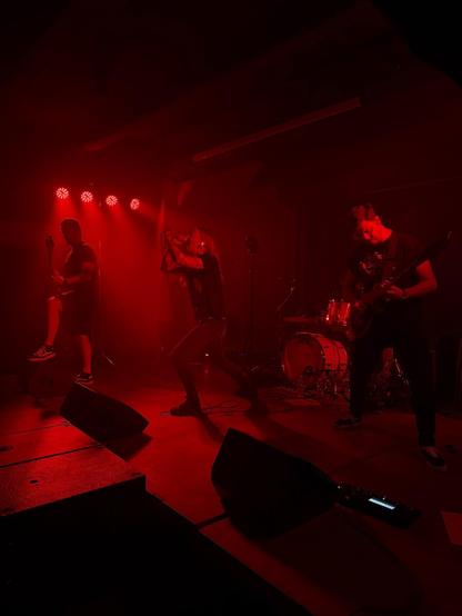 Band Kalypso on stage. Drenched in red light. Three band members playing in front of the drum kit.
