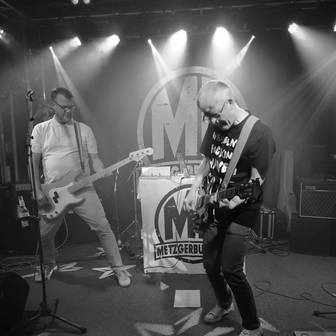 Zwei Männer stehen auf einer Bühne und spielen Gitarre und Bassgitarre. Zwischen ihnen steht ein Tisch, an dem ein Banner mit der Aufschrift "MB MetzgerButcher" hängt.