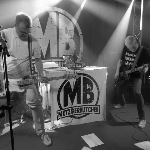Zwei Männer stehen auf einer Bühne und spielen Gitarre und Bassgitarre. Zwischen ihnen steht ein Tisch, an dem ein Banner mit der Aufschrift "MB MetzgerButcher" hängt.