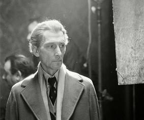 This is a photo of Peter Cushing with lighter, whiter hair. He's looking off-screen, and wearing an overcoat, scarf, and a three-piece suit and tie.