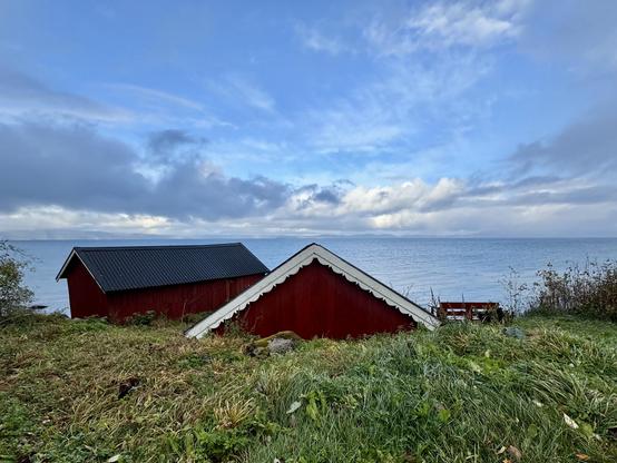 The apex of a red house is visible above a foreground of green grass, its white gingerbread eaves barely touching the horizon dividing blue sea and sky in the background.