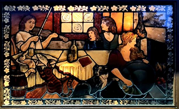 A stained glass window depicting a Highland scene of women working in a home while listening to someone playing a fiddle.