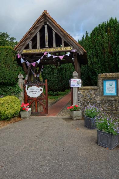 A quaint, rustic wooden gatehouse, reminiscent of a traditional English lodge, stands at the entrance of a garden or churchyard. The gatehouse features a steep, tiled roof with exposed wooden beams and a sign above reading, "Blessed are the Dead which die in the LORD." The entrance is adorned with a decorative bunting of pink, white, and purple triangular flags. A wooden gate, slightly ajar, displays a sign that reads, "Cream Teas Shoreham Church Today," accompanied by a teapot illustration. To the right of the gate, a signpost indicates that cream teas are available today. The surroundings are lush with greenery, including neatly trimmed bushes and potted flowers in vibrant colours, adding a welcoming touch to the scene. The pathway leading through the gate is paved with red bricks, inviting visitors to explore further.