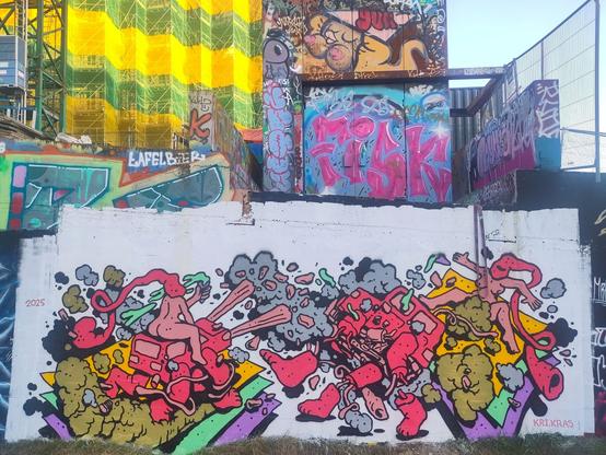 Photo of a graffiti piece, taking up the bottom half of the picture, on a concrete wall, about 4m high, 8-10m wide. White background, cartoonesque figures painted on it. Two red cars on four feet, each ridden by a naked female human character, heads wrapped in red cloth, hanging behind them. The cars seem to attack each other, with sort of stone rays coming from the headlights, and the car on the right falling apart, surrounded by grey dust. The character on the right seems to fall of on three stacked sheets, yellow, green, purple from top to bottom. Same sheets are below the car on the left.
In the bottom-right corner a tag that says "KRI.KRAS", in the top-left corner the year "2025".

The top of the picture is several graffiti works, that have been here for quite a while, among others a female Utrecht gnome ("Utrechtse kabouter"), with long blonde hair, and large bare breasts. In the background, top-left, a building surrounded by scaffolding wrapped in green and yellow netting.