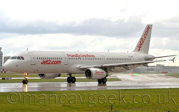Side view of a twin engined jet airliner parked on a runway, ready for take-off.
The plane is basically white, with red "Jet2.com" titles on the lower forward fuselage, red "Friendly Low Fares" text on the upper middle fuselage, over the wings, and both sets of text on the white engine pods under the wings.
There are also diagonal red "Jet2.com" title on the white tail.
Several lights on the nose-wheel leg are reflected on the damp runway.
Grass fills the foreground, with more grass visible under the plane in the background, leading up to large terminal buildings in the distance on both sides of the frame.
Bright but cloudy sky completes the image.