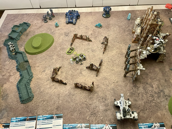 A Warhammer battle on a table, seen from above. Terrain pieces, particularly ruins. Ultramarines company heroes, a land raider and a ballistus dreadnought on one side of the table. Two t’au stealth battlesuit teams and a ghostkeel battlesuit deployed forward, as well as a pathfinder team hidden in the ruins. A hammerhead tank behind.