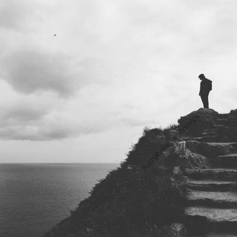 Black-and-white photograph of a person standing alone at the edge of a cliff at Binn Éadair (Howth), Ireland. The sea stretches beneath a heavy sky, and the rocky steps lead into the horizon — a scene suspended between solitude and reflection.