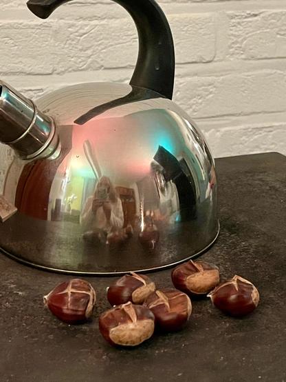 Chestnuts slowly roasting on a stove, next to a stainless steel kettle that reflects the living room around it (and me holding my phone). A white painted brick wall in the background.