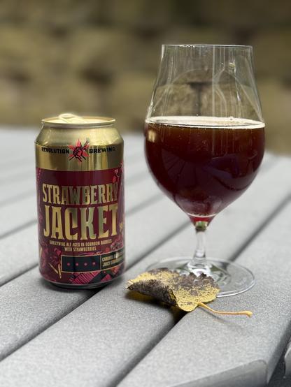 A shiny gold can with a deep red label and gold lettering sits on a gray picnic table next to a snifter glass full of the deep red-brown liquid. A fallen leaf is yellow with a speckled brown background