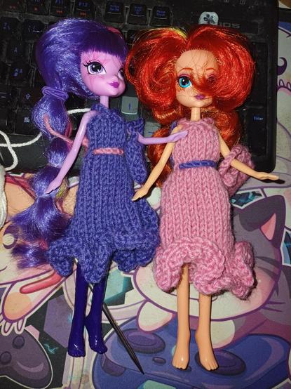 Kaksi kirkasväristä muotinukkea yllään neulotut röyhelöhelmaiset mekot, toisella pinkki violetilla vyöllä, toisella violetti pinkillä vyöllä.