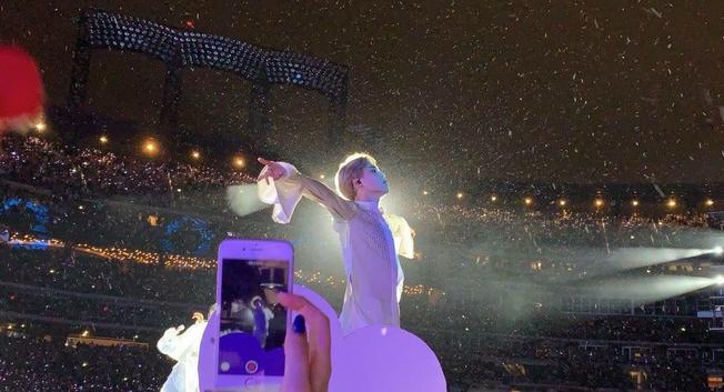 A photo of Jimin of BTS, holding his arms out wide and backlit by the concert lights.