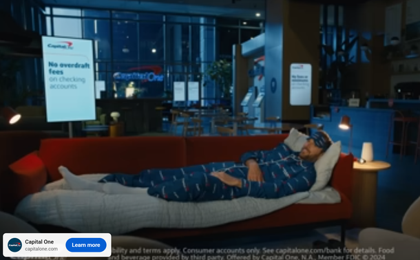 Screen grab of an online ad for a credit card company. The spokesperson/ character who's selling the bank's services is seen in pyjamas, on a blanketed sofa in the lobby of a hotel branch office. He's about to go to sleep, supposedly. A wall of windows in the background show faded light, perhaps clouds or early evening.