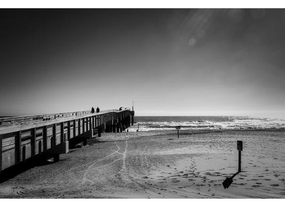 Black and white photo of a wooden pier stretching out to a calm ocean, with two silhouetted figures walking. Empty sandy beach in the foreground. Tranquil mood.