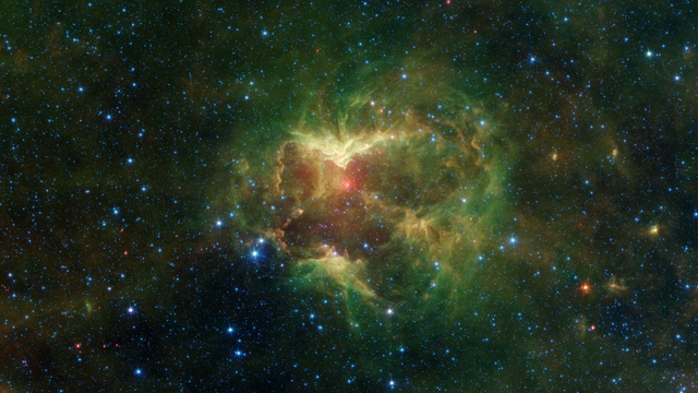 Infrared image of the Jack-o’-Lantern Nebula captured by NASA’s Spitzer Space Telescope, showing a glowing cloud of green and orange gas and dust with a bright red center shaped like a carved pumpkin, surrounded by a field of blue stars scattered across space.