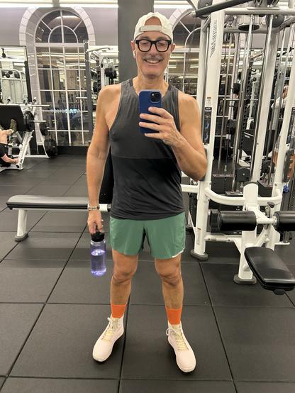 A man (me) is smiling and taking a selfie in a gym. He is wearing a sleeveless black top, green shorts, and light pink shoes with orange socks. He holds a water bottle in one hand and is wearing glasses and earbuds.