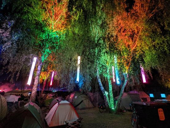 Einige Zelte unter Bäumen, die farbstark in allen Farben des Spektrums beleuchtet werden. Nachts aufgenommen ist das Blattwerk farbig, doch blendfrei, ohne Überstrahlungen und nicht übermäßig hell.