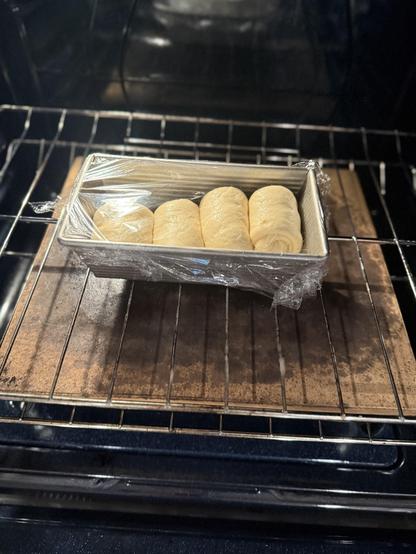 A loaf pan with rising dough rolls covered in plastic wrap is placed in an oven.