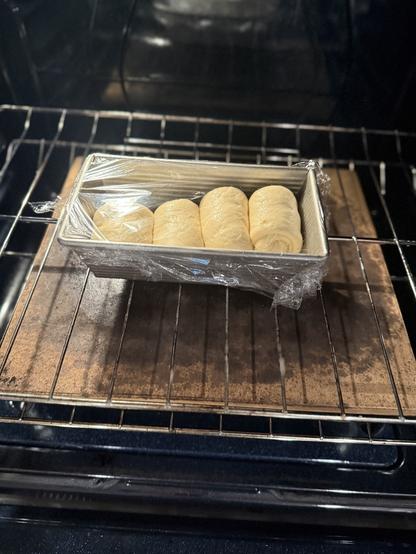 A loaf pan with rising dough rolls covered in plastic wrap is placed in an oven.