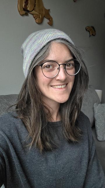 A woman with brown hair and black round glasses is looking at the camera and smiling. She is wearing a gray beanie hat.