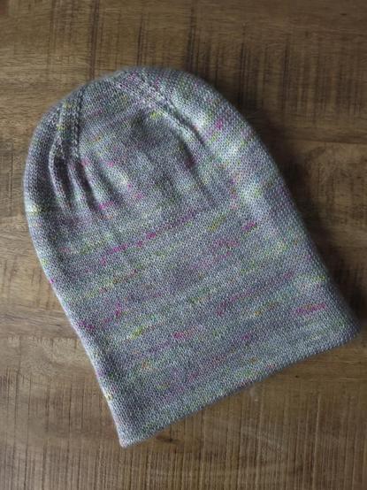 A double-layered hat is laid flat on a wooden surface. The hat is knit with handyed yarn in gray with neon yellow and pink speckles.