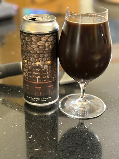 A silver pint can has a label with an image of a bar with walls and ceiling covered with plates. The snifter glass to the right is very full of dark brown beer