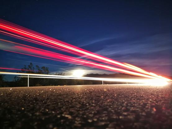The image shows a low-angle view of a road at night with the moon rising over distant mountains and scattered clouds in a dark blue, star dotted sky. Red and white vehicular lights streak across the scene.